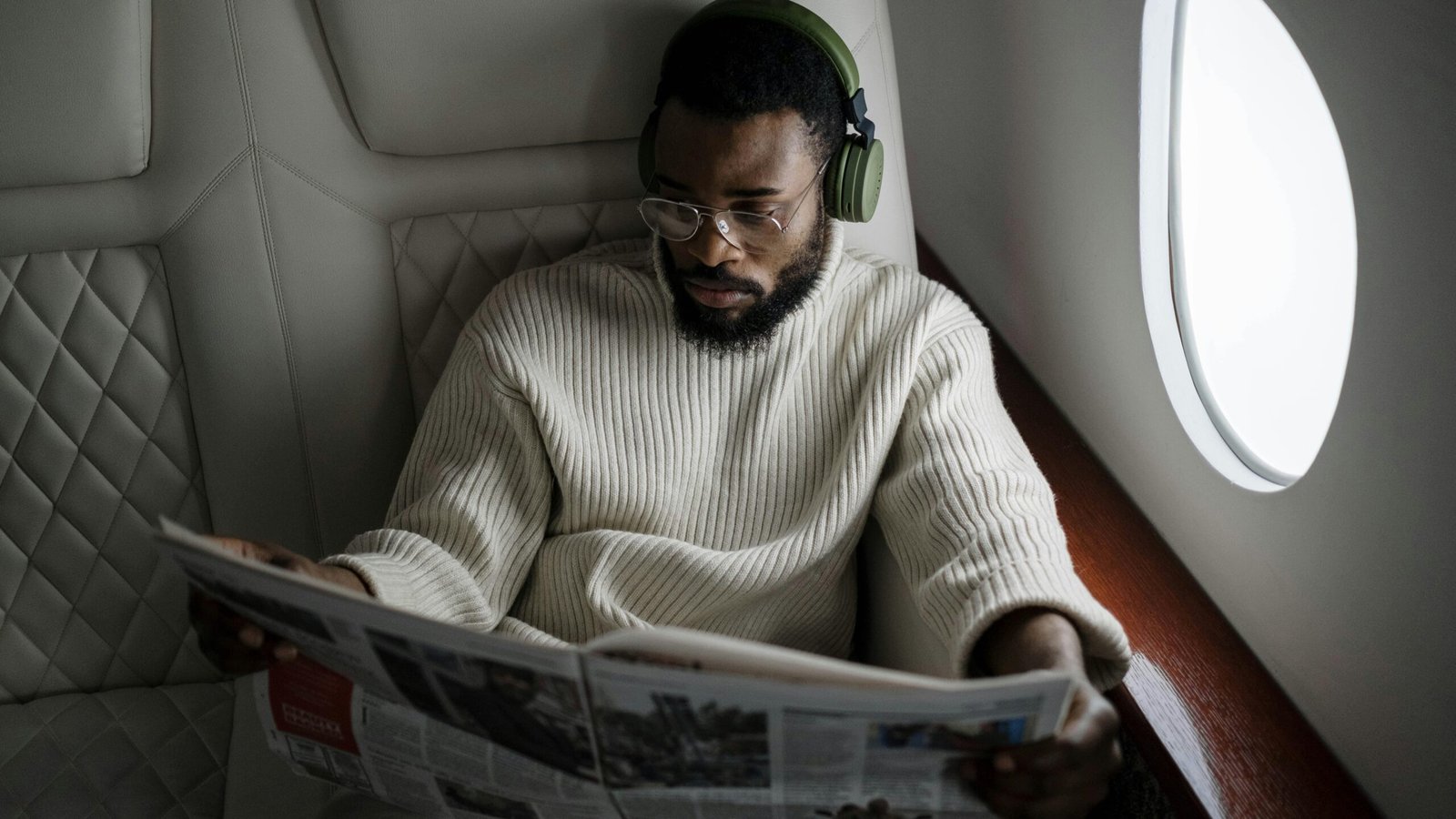 Business man reading newspaper on airplane wearing headphones and glasses.