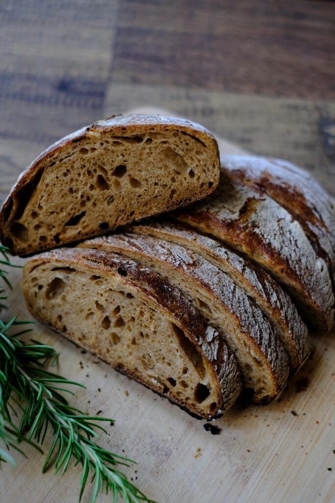 Close-up of a sliced, crusty sourdough loaf with fresh rosemary on a wooden surface.